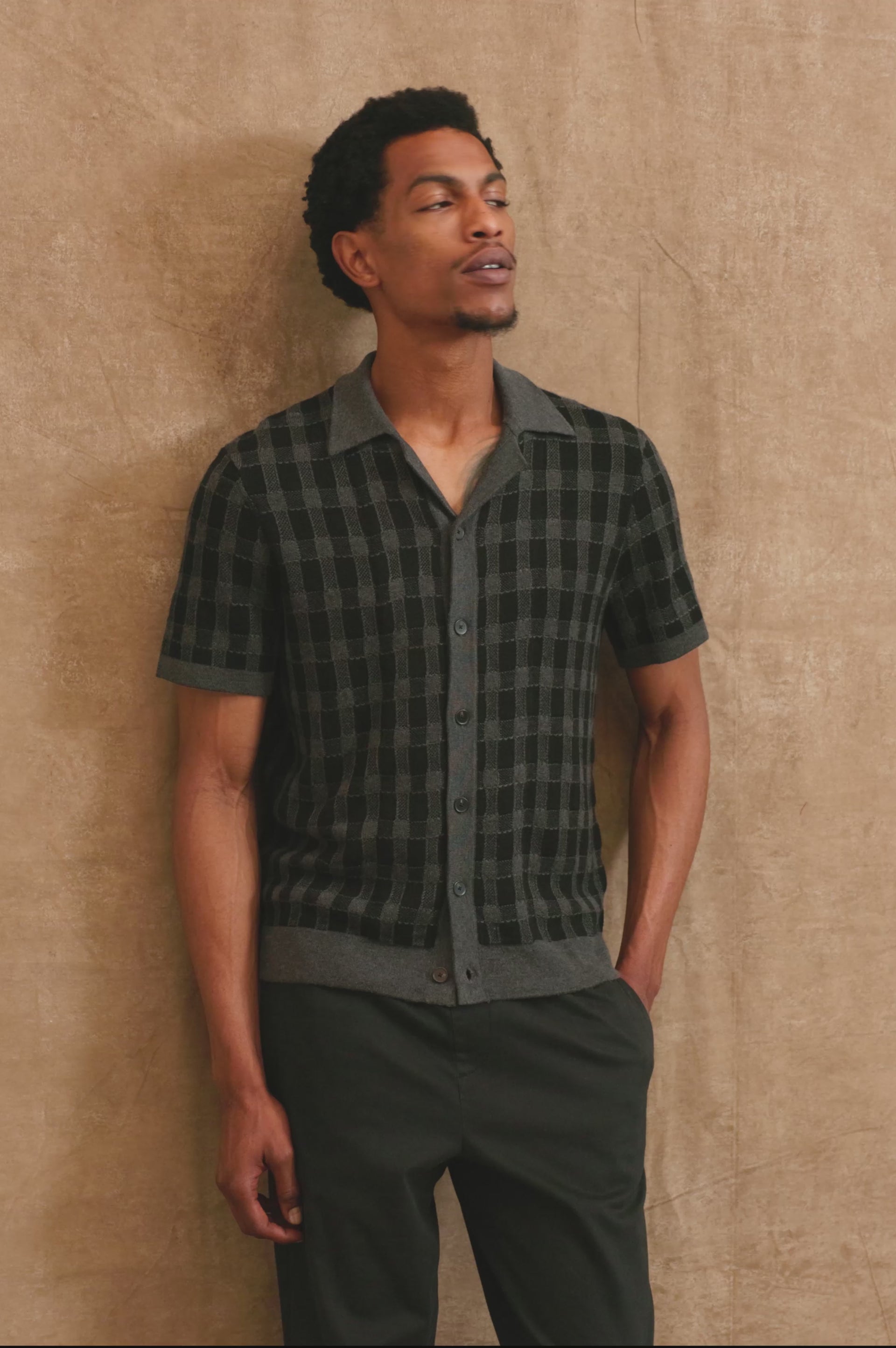 The Ronan is a collared button front knit shirt with short sleeves, grey rib trim, grey + black block stitch pattern in Charcoal Black colorway. Front view video with sunglasses featured on model.