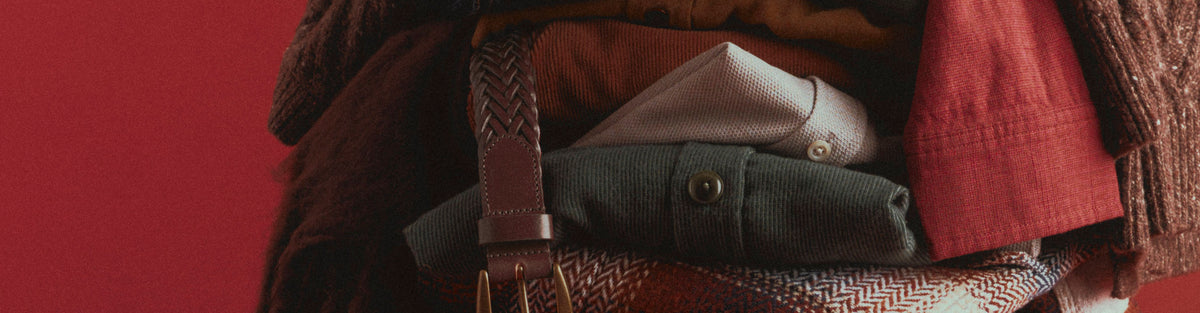 A close-up of folded clothing and a patterned herringbone bag against a solid red background. Visible items include a dark green corduroy jacket, a light gray shirt, a rust-red fabric piece, and a brown woven strap.