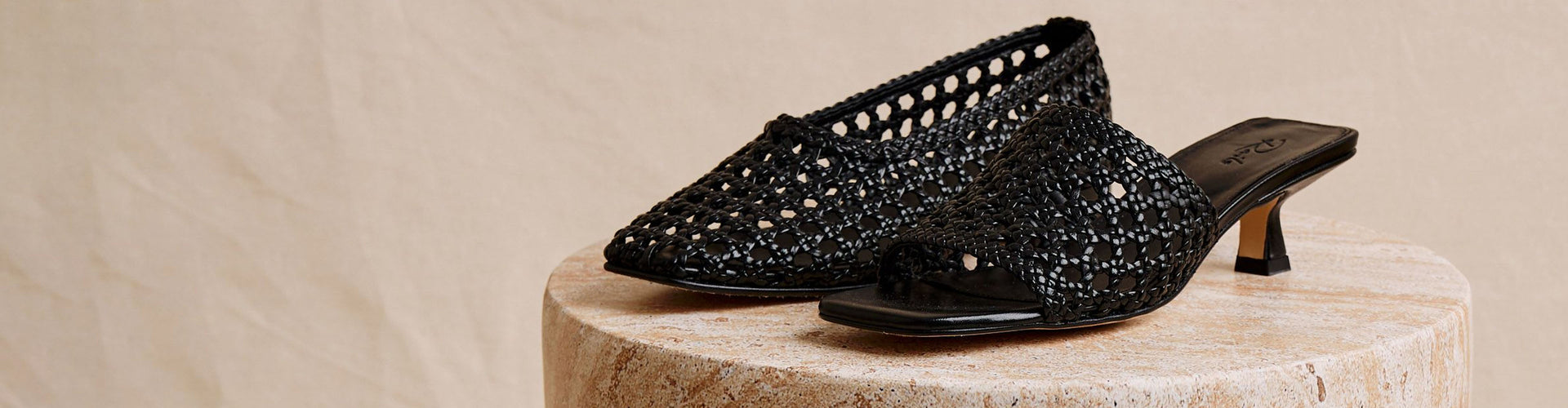 A pair of black woven leather kitten-heel mules with an open square toe, displayed on a round stone pedestal against a neutral beige background.