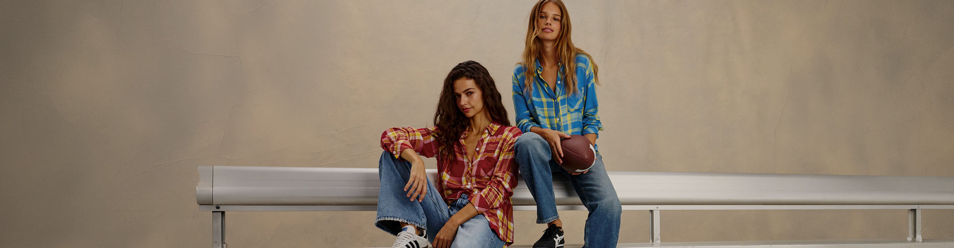 Two women sitting on a metal bench wearing matching patterned long-sleeve shirts and blue jeans, posed against a neutral beige studio background.
