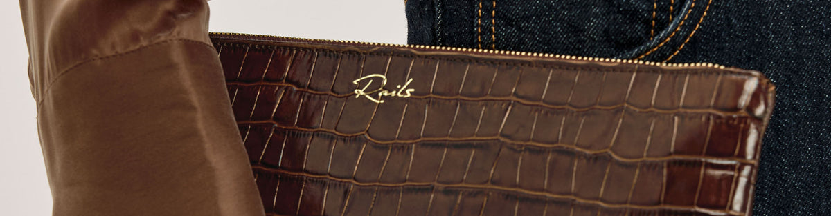 A close-up of a brown crocodile-embossed leather clutch or pouch with a gold script logo reading 'Rav1s' on the front, shown against a neutral background with part of a brown sleeve and dark denim visible.