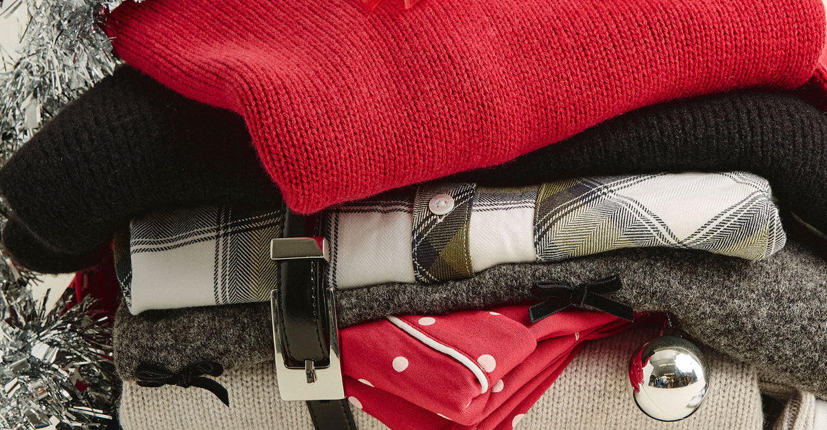 A close-up stack of folded knit and woven clothing items, including a bright red sweater, a black knit garment, plaid shirts in white and gray, a gray knit piece, and a red fabric item with white polka dots, arranged together with a silver ornament.