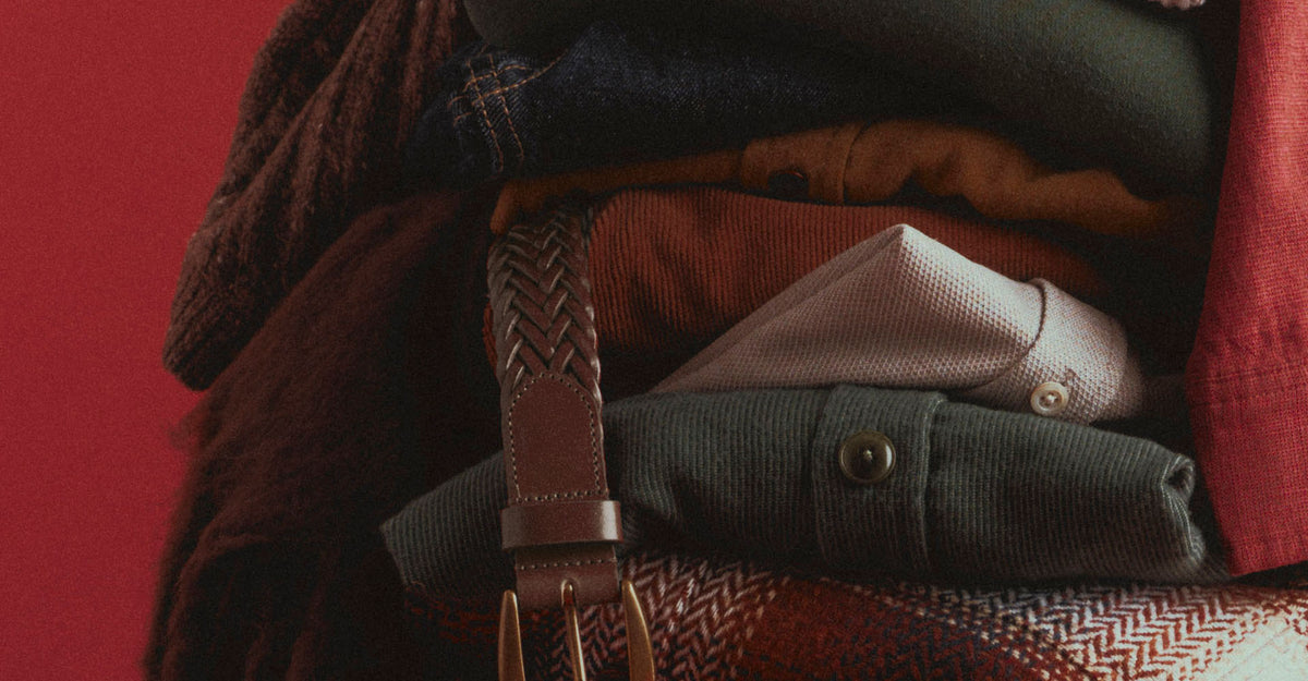 A close-up of a styled stack of folded clothing and accessories against a solid red background, featuring a brown braided belt with gold-toned metal hardware draped over a dark green corduroy shirt and other garments.