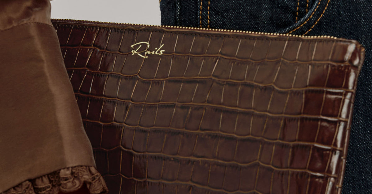 A close-up of a brown crocodile-embossed leather clutch or pouch with a gold script logo reading 'Rivis' on the front, shown next to a brown satin fabric and dark blue denim.