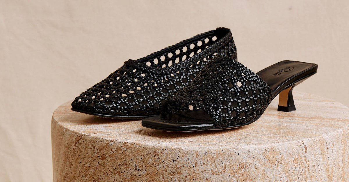 A pair of black woven leather kitten-heel mules with an open square toe, displayed on a round stone pedestal against a neutral beige background.