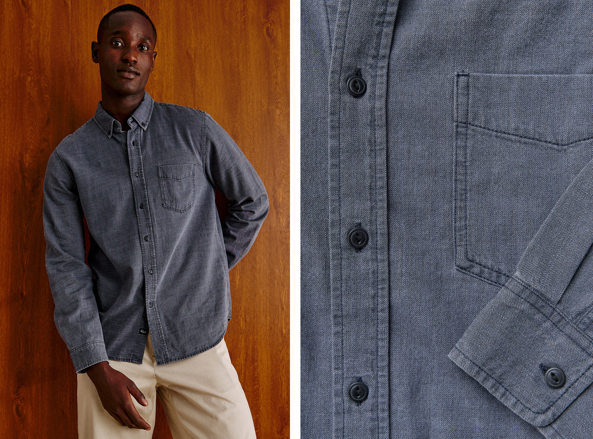 A man wearing a slate blue button-down shirt with beige pants, standing against a warm wood-paneled wall, with a close-up inset of the shirt's chest pocket and button details on the right.