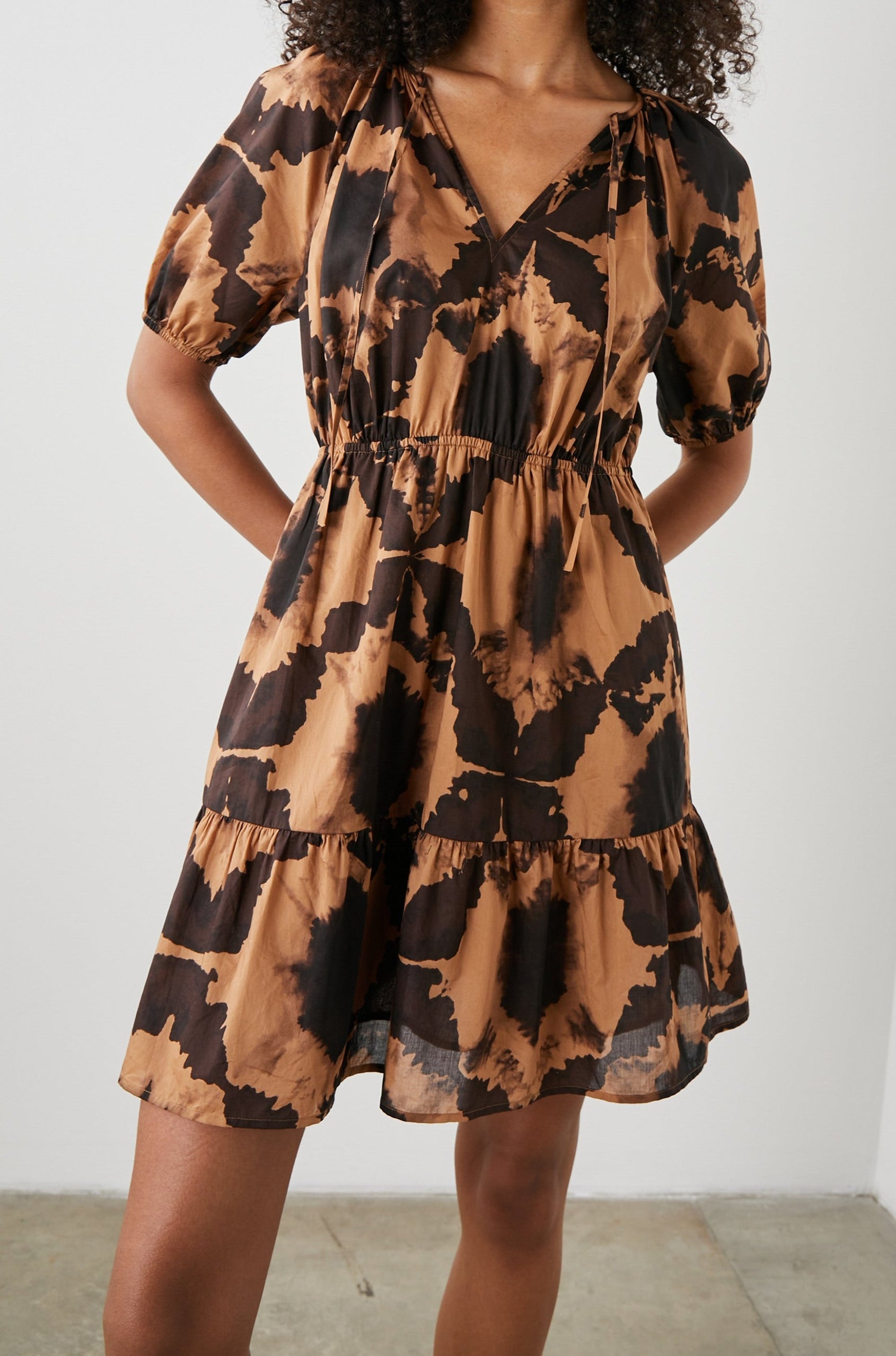 Amabella Short Sleeve Mini Dress with a V neck with Ties, Puff Sleeves, a Cinched Waist, and a Tiered Skirt in Tigers Eye Colorway, a Floral Pattern in Browns on Dark Brown - Front View Featured on Model  